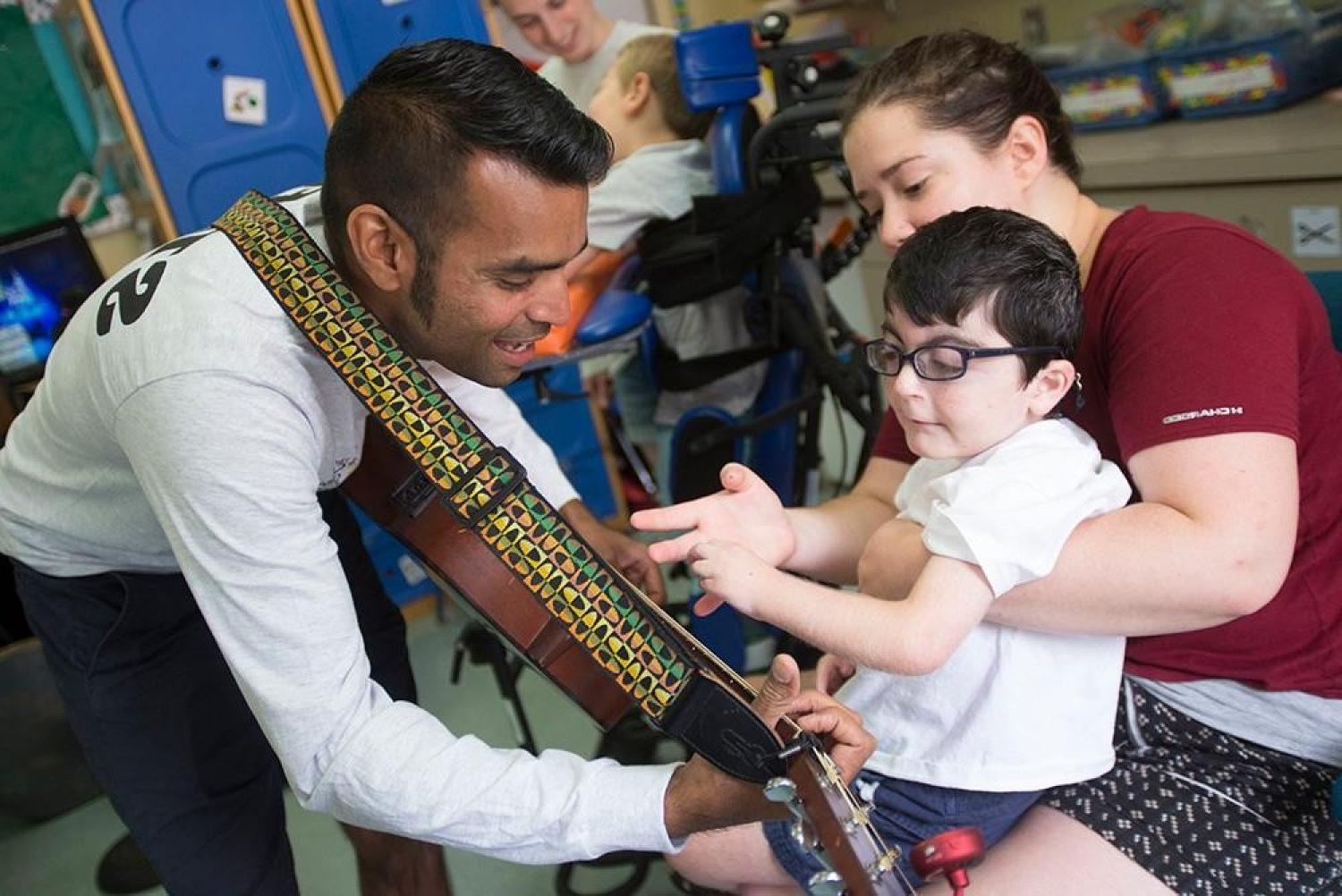 A young boy sitting on a teacher's lap watching a man play guitar