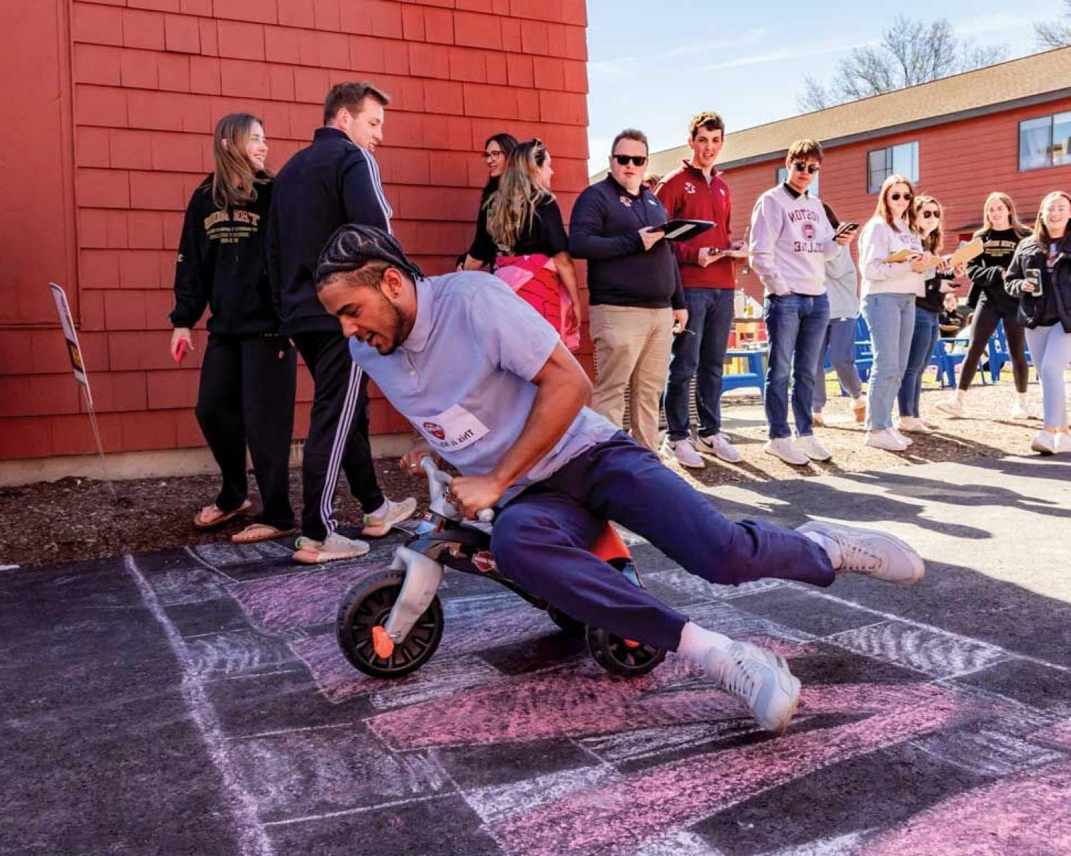 Miles Hester ’24 crosses a chalk drawn finish line on a trike with fellow student onlookers.